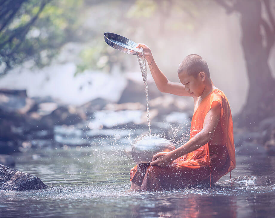 Very young Buddhist monk sitting in river washing cooking pot