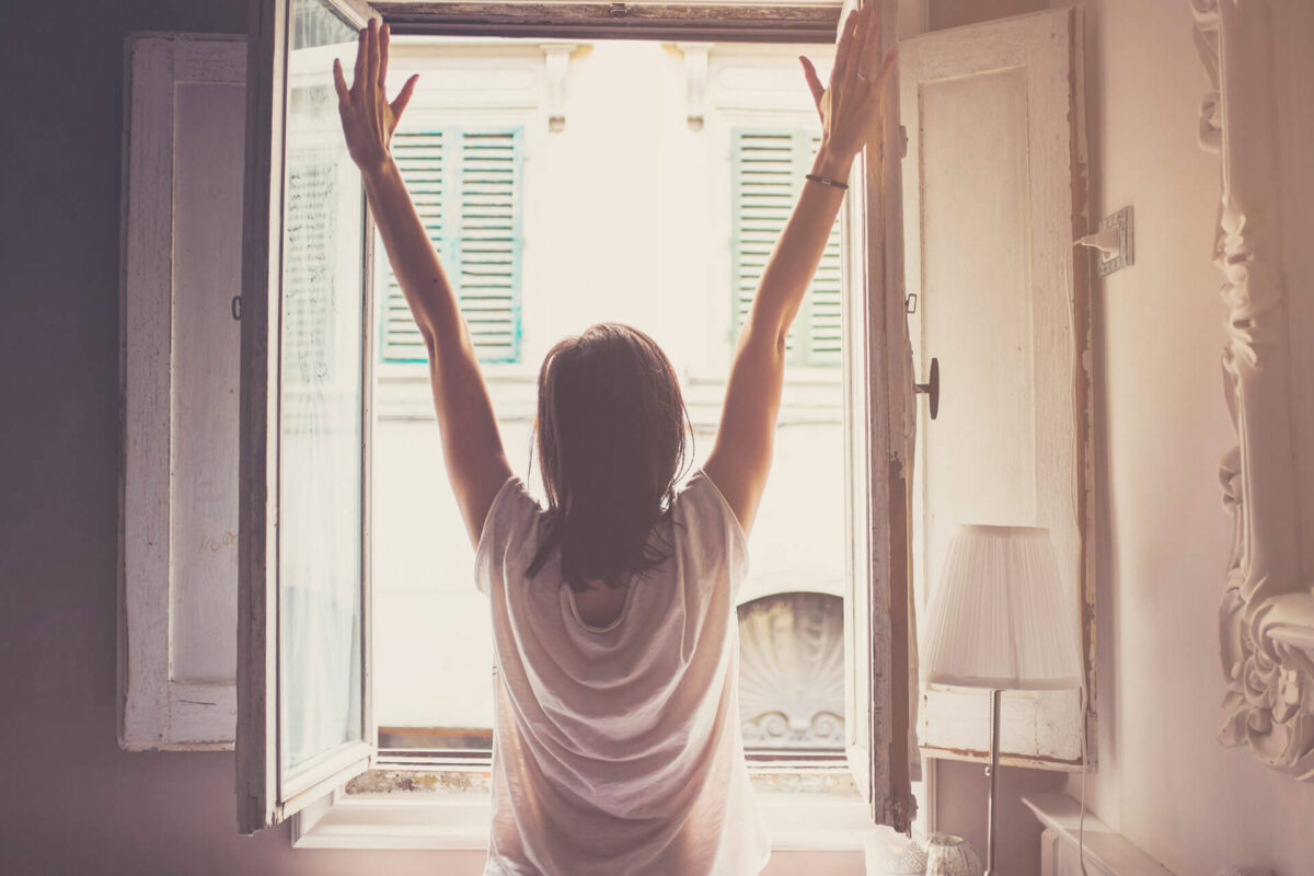 Mujer estira las manos hacia arriba, fotografiada de espaldas frente a una ventana abierta