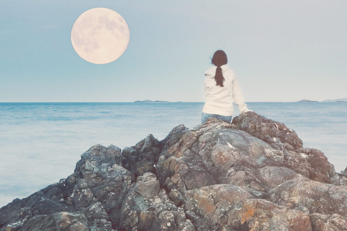 Mujer fotografiada de espaldas, sentada en las rocas de la playa y observando la luna llena durante el día